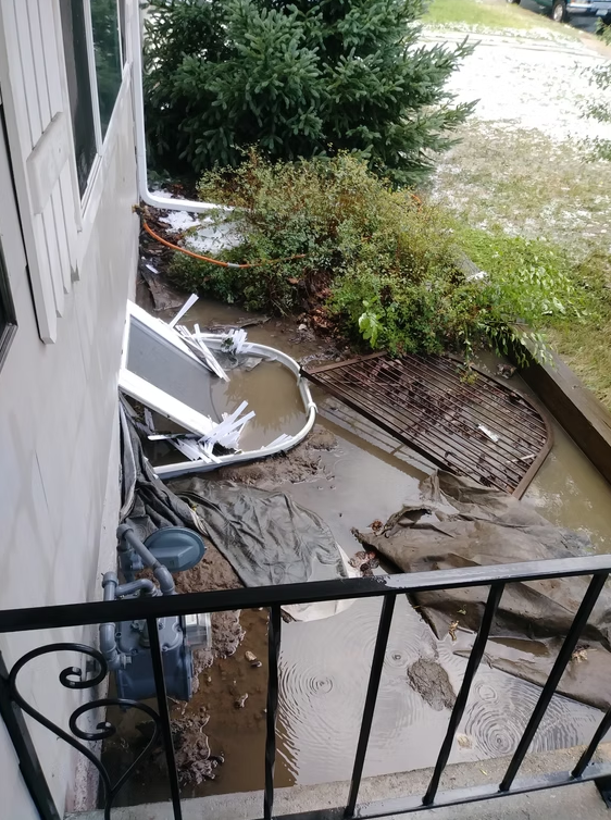 Broken fence, muddy water, and debris scattered near the corner of a house. Damaged metal and plastic pieces are visible by bushes, with water pooling on the ground after apparent flooding or a storm.