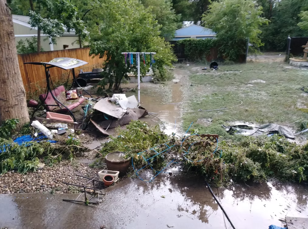 A backyard is flooded and cluttered with debris, broken furniture, fallen branches, and scattered objects. The grass is wet and muddy, and water covers parts of the ground. Trees and a wooden fence are visible in the background.