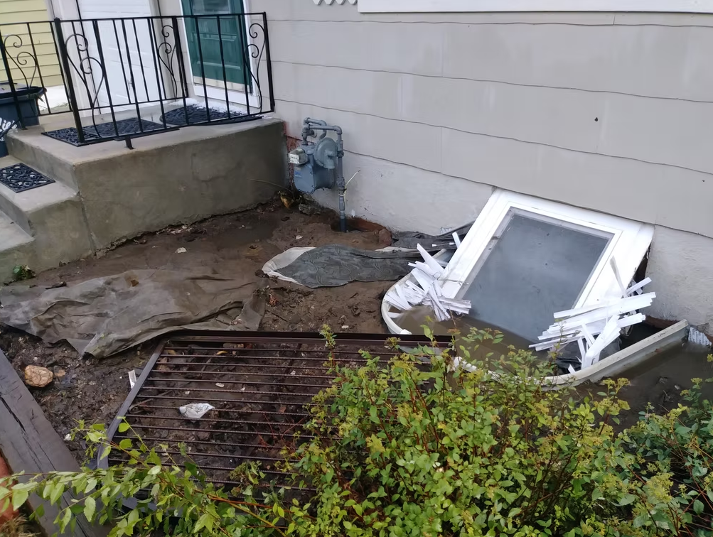 A damaged basement window well with debris, a broken window, and torn blinds sits beside a house. Mud and tarp cover the ground, and a metal grate leans nearby. A bush is in the foreground near the concrete steps.