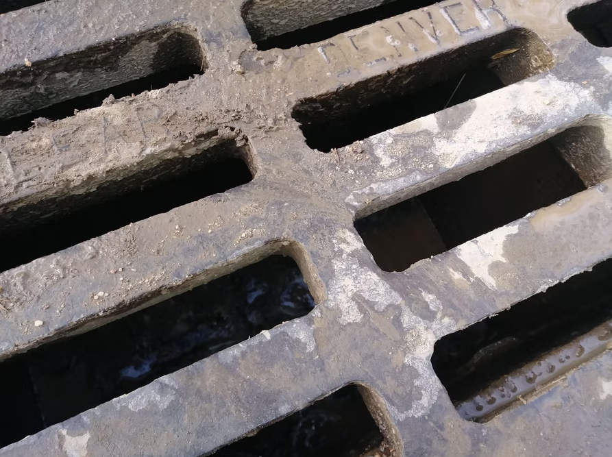 A close-up view of a dirty, weathered metal sewer grate with the word SEWER partially visible on the top right corner. The surface shows signs of rust and grime buildup.