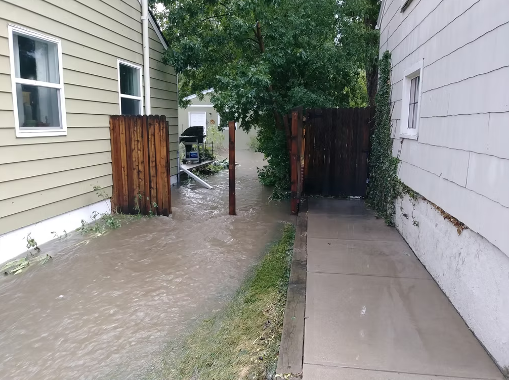 Floodwater flows between two houses and through a partially open wooden gate, covering the yard and sidewalk. Trees and a garage are visible in the background, and the sky appears overcast.