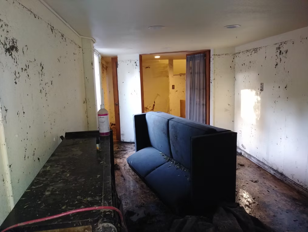 A dimly lit room with dirty, stained walls and floor. A large, overturned blue couch sits in the center, and debris is scattered around. The space looks abandoned and damaged, possibly by flooding or neglect.