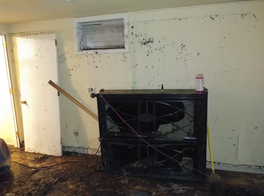 A basement room with dirty, mud-stained walls and floor, open white door, an air vent, overturned shelving, a broom, and a large bottle sit among debris after apparent flooding.
