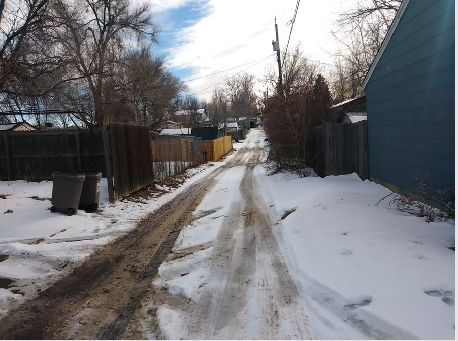 A narrow, snow-covered alley with muddy tire tracks runs between fences and houses on a winter day. Trash bins are on the left, and bare trees line the sides under a partly cloudy sky.