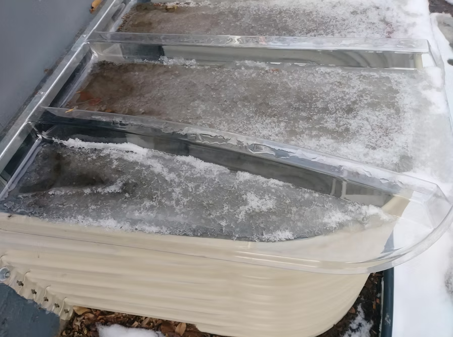 A hot tub covered with a thick layer of ice and snow, with clear frozen water forming ridges on top of its cover. Some leaves are visible beneath the icy surface.