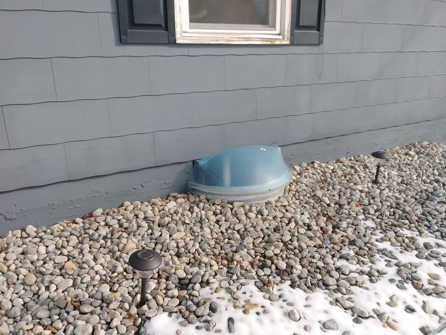 A gray house exterior with a window above a plastic window well cover surrounded by rocks and patches of snow. Two small outdoor lights are visible among the rocks.