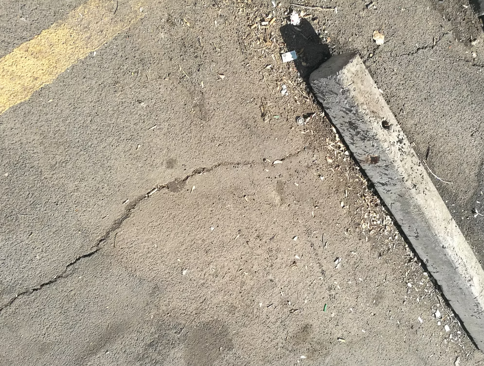 A cracked asphalt parking lot with a concrete parking block, scattered debris, and a faded yellow line partially visible on the upper left.