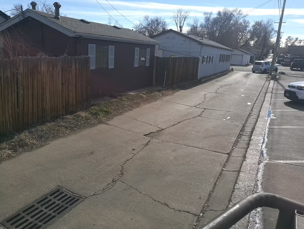 A cracked concrete alley runs between a wooden fence and buildings on a sunny day. Parked cars are visible on the right side, and leafless trees stand in the background.