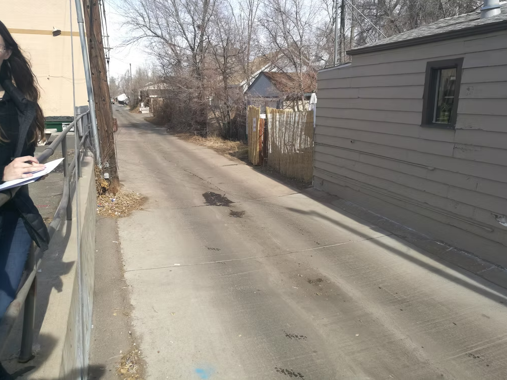 A narrow alleyway with bare trees and houses on each side. A person on the left, partly out of frame, is holding a notepad and pen, standing by a metal railing. Shadows are cast across the concrete path.