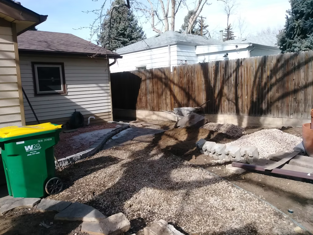 A backyard with a wooden fence, gravel pathways, a green Waste Management trash bin, a tan house, and a partially landscaped area with dirt, rocks, and some potted plants. Trees cast shadows across the yard.