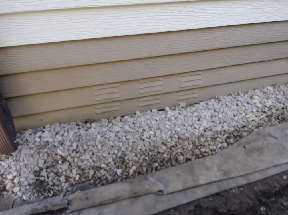White rocks cover the ground next to the beige siding of a house. Multiple rectangular vents are visible on the wall above the rocks, and a concrete or stone edge borders the rocks on one side.