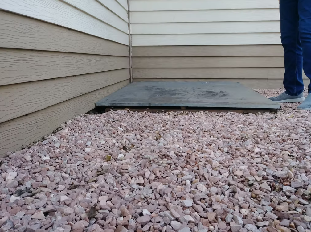 A person wearing jeans and gray shoes stands near a concrete slab at the corner of a building with tan siding, surrounded by pinkish gravel stones.