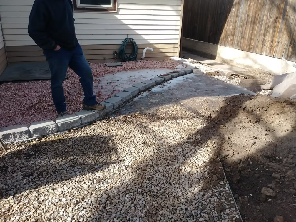 A person stands on a gravel area next to a house with beige siding, near a row of gray bricks marking the edge of a walkway. A garden hose and wooden fence are visible in the background.