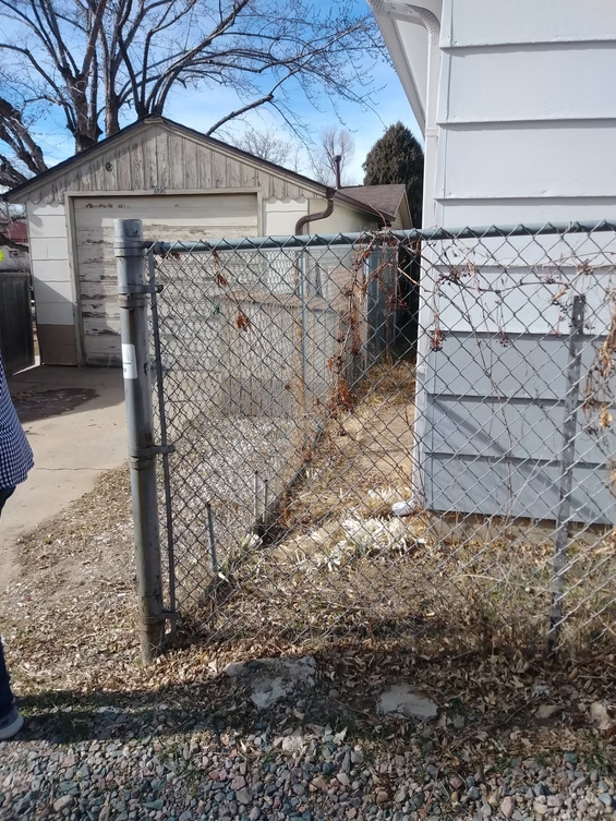 A chain-link fence with a gate is next to a white house, leading to a narrow, gravel-filled side yard. In the background, there is a weathered wooden garage and leafless trees.