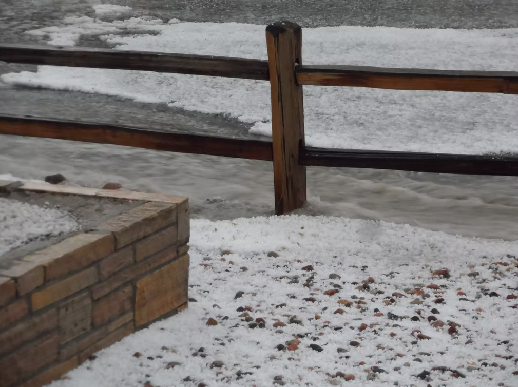 A wooden fence stands beside a flooded area; the ground, river, and nearby rocks are all covered with a thick layer of hailstones.