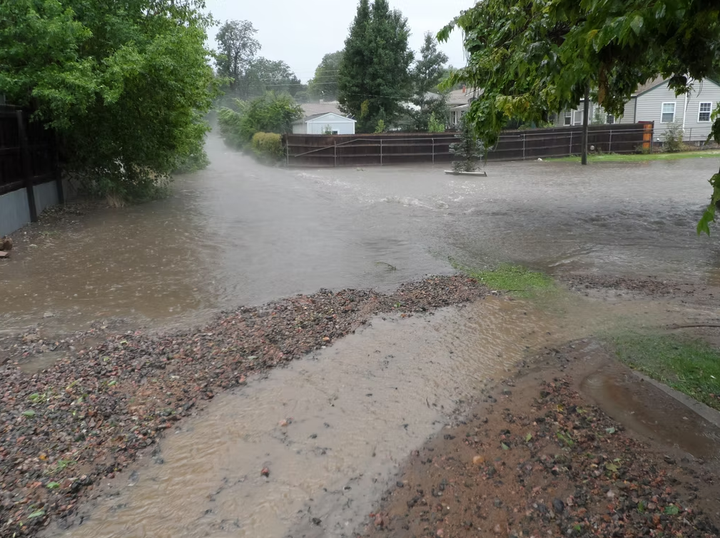 A residential area is flooded after heavy rain, with water covering the street, driveway, and yards. Trees and houses are visible in the background under an overcast sky.