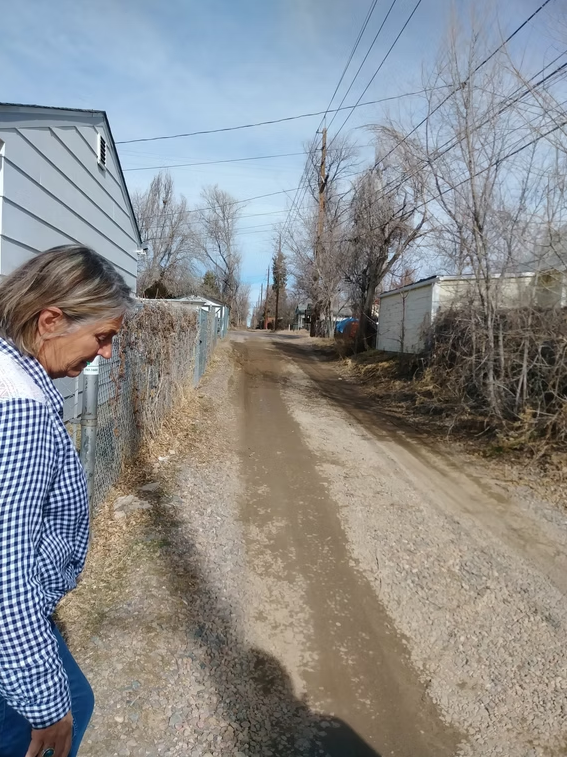 An older person with gray hair, wearing a blue and white checkered shirt, walks beside a dirt alley bordered by fences, leafless trees, and small houses on a clear day.
