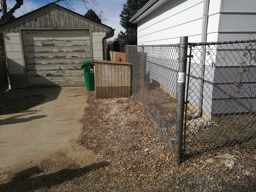 A weathered wooden garage sits beside a white house. A green trash bin is near the garage, and a small wooden shed stands between the buildings. A chain-link fence runs along the right side of the scene.