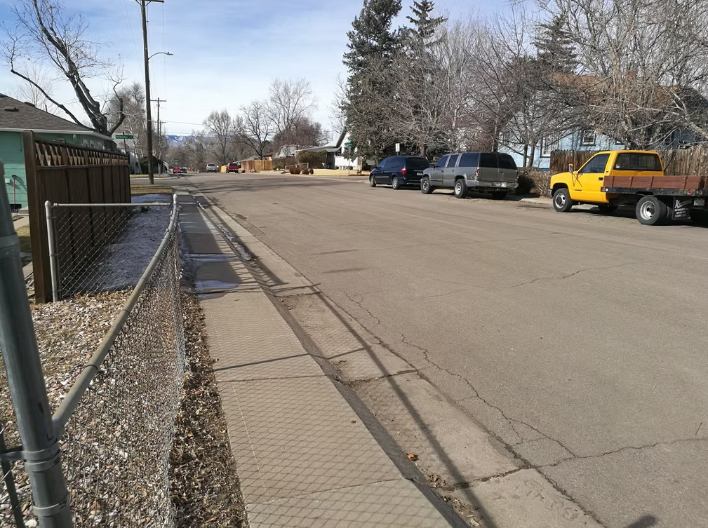 A quiet suburban street with several parked cars, including a yellow truck, lined by leafless trees and houses. A chain-link fence runs along the sidewalk on the left side of the image.