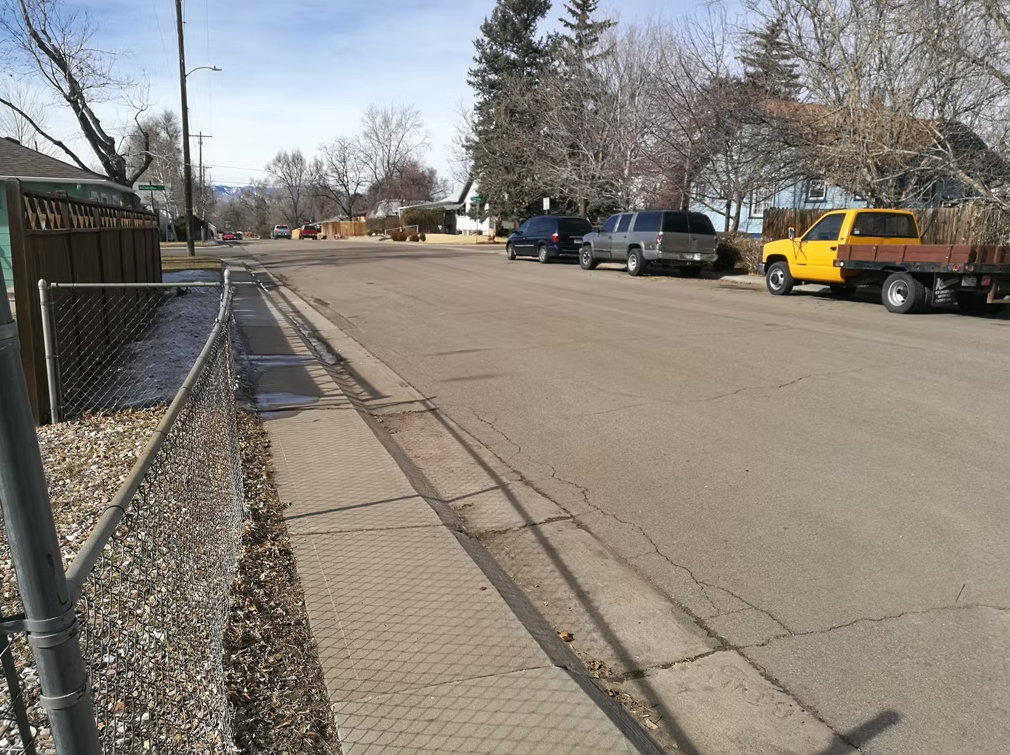 A quiet suburban street with several parked vehicles, including a yellow pickup truck, runs alongside a chain-link fence, trees without leaves, and houses on a clear day.