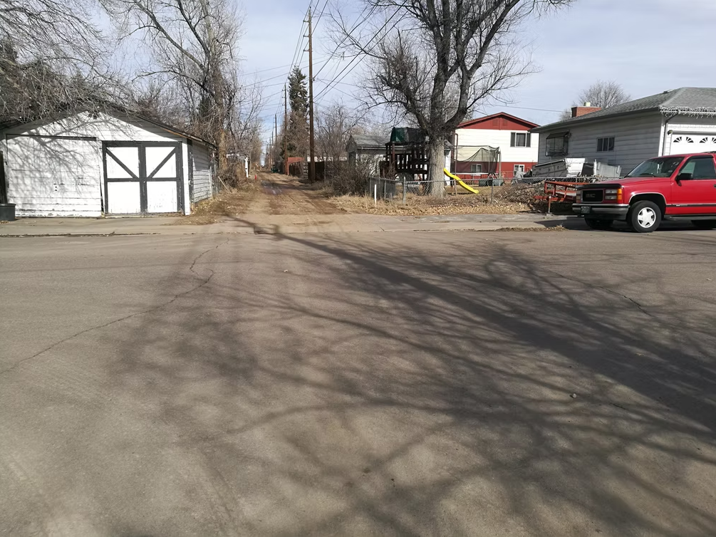 A paved street meets a dirt alley between houses. A white garage is on the left, a red truck is parked on the right, and there are leafless trees casting shadows across the road.