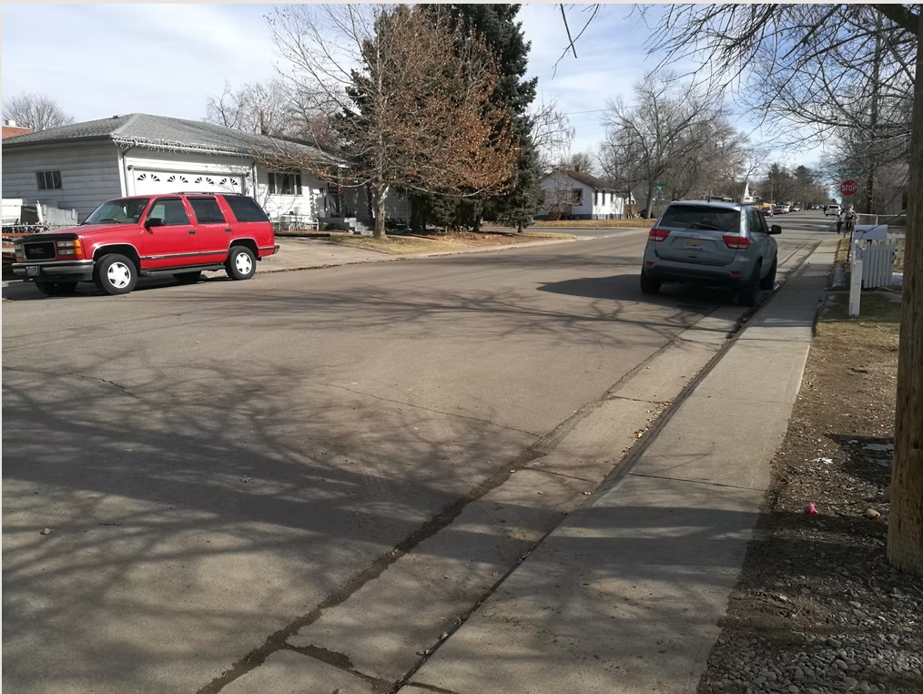 A quiet suburban street with a red pickup truck parked on the left side and a gray SUV parked on the right. Leafless trees line the sidewalk, and houses are visible in the background on a clear day.