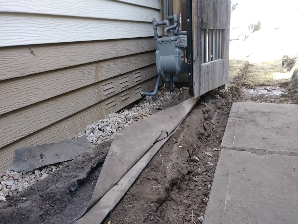 A utility meter is mounted on the side of a tan house near a wooden fence. The ground is excavated, exposing soil and rocks, with pieces of fabric or tarp laid out beside the trench. Concrete pavement is on the right.