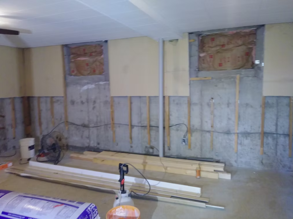 An unfinished basement with exposed concrete walls, insulation, wooden studs, and construction materials such as lumber and tools scattered on the floor. Some electrical cords and equipment are also visible.