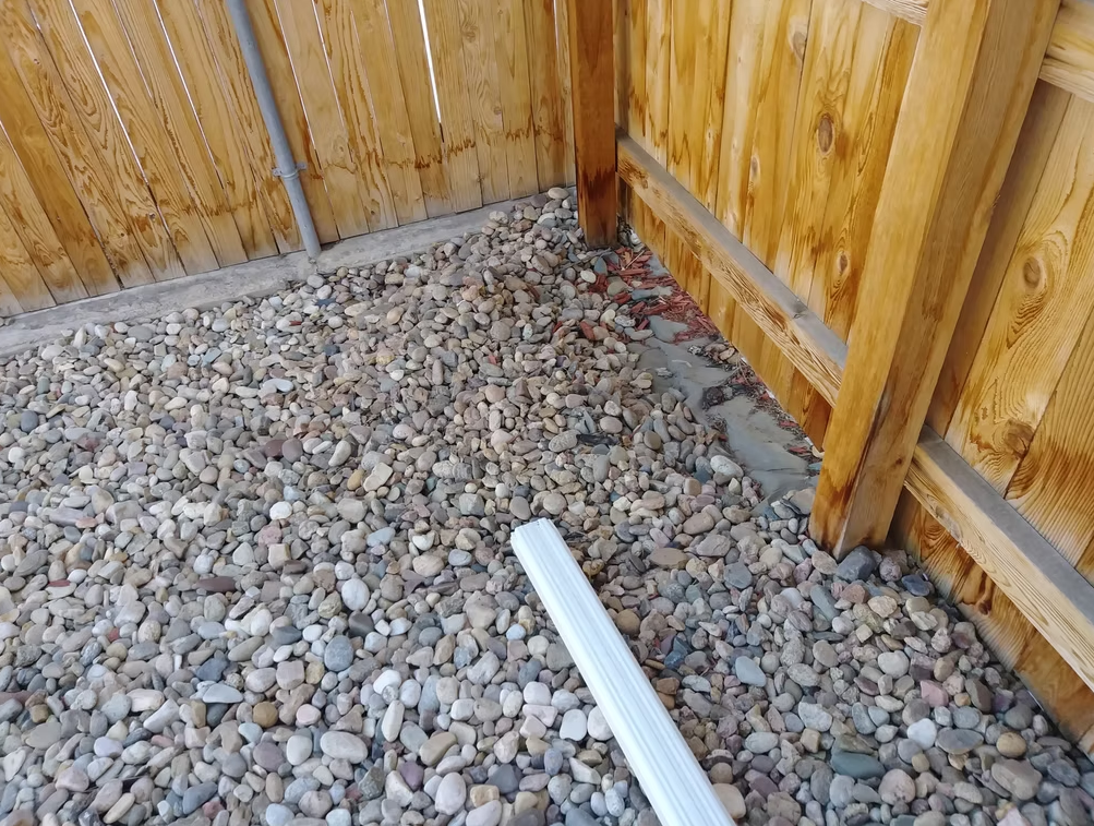 A section of a backyard with a wooden fence and rocky ground. A white plastic gutter downspout extension lies on the rocks near the fence. Some red mulch is visible in the corner by the fence.