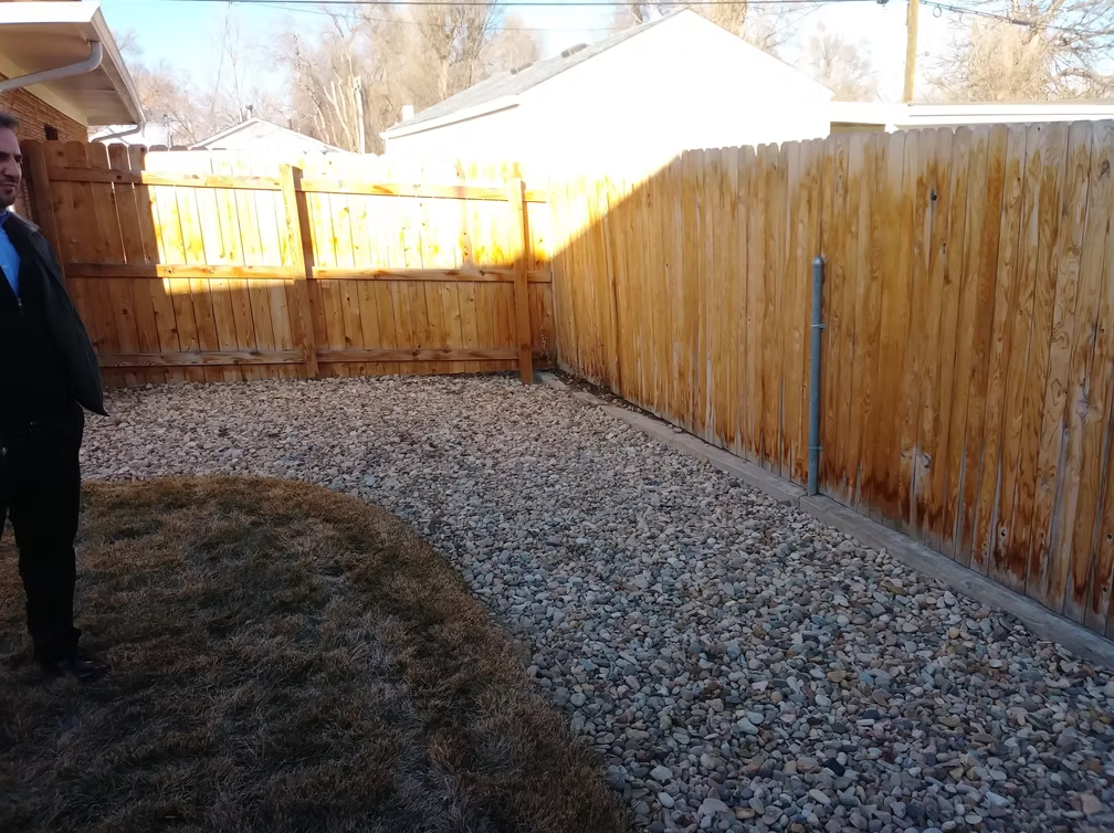 A man in a black jacket stands on the edge of a small backyard with a wooden fence and a section of grass bordering an area covered with rocks. Sheds and leafless trees are visible in the background.