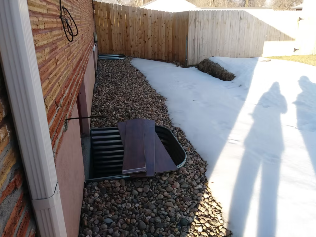A house with brick siding has basement window wells, one covered by wood planks. The ground is covered in rocks near the building and snow further out. Two people’s shadows are visible on the snow by a wooden fence.