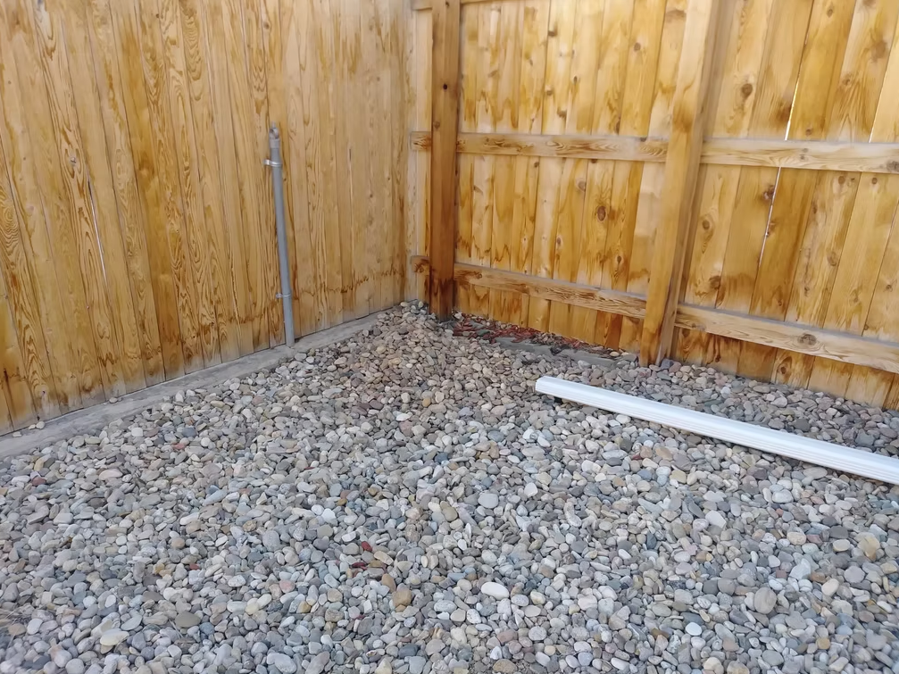 A corner of a backyard with a wooden fence, gravel covering the ground, a vertical metal pipe on the left, and a white rectangular object lying on the rocks to the right.