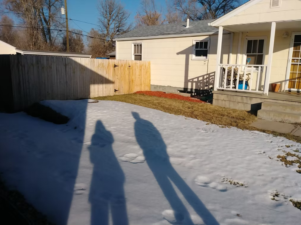 Two tall shadows of people stretch across a snow-covered yard toward a yellow house with a small porch, a fenced backyard, and bare trees in the background under a clear blue sky.