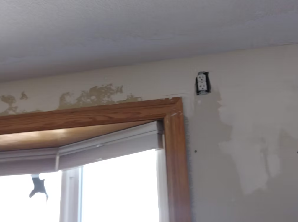 A partially renovated room with unfinished drywall, a window with wooden trim, and a broken window blind. An electrical outlet without a cover is visible near the ceiling.