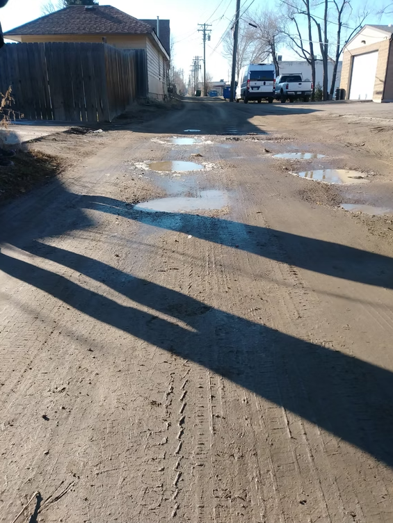 A dirt alleyway with several puddles, deep tire tracks, and long shadows cast by people. In the background, there are houses, a wooden fence, two white vans, and leafless trees.