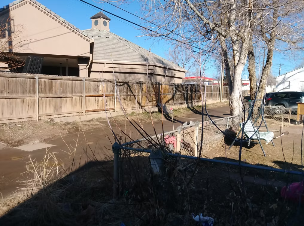 A backyard with bare trees, a chain-link fence, a swinging bench, and a dirt alleyway beside a wooden fence. A house and parked car are visible under a clear blue sky.