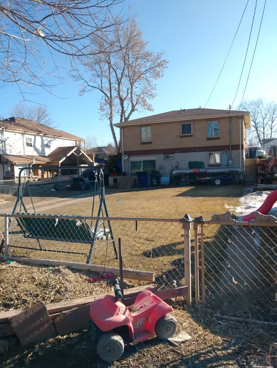 A fenced backyard with a swing set, a red toy car, and scattered objects. The yard has patchy grass, and a two-story house stands in the background under a clear blue sky with bare trees.