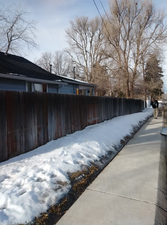 A sidewalk runs alongside a wooden fence with patches of melting snow on the ground. Bare trees stand in the background, and a person wearing a dark jacket and khaki pants is walking toward the camera.
