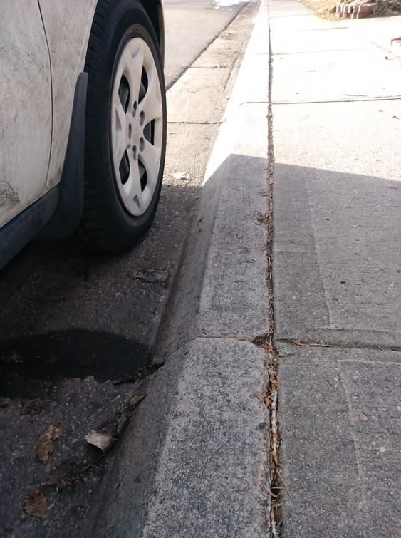 A close-up of a car’s front wheel parked next to a curb on a sunny day. The curb and sidewalk are visible, with some dirt, leaves, and small debris along the edge. Shadows stretch across the pavement.