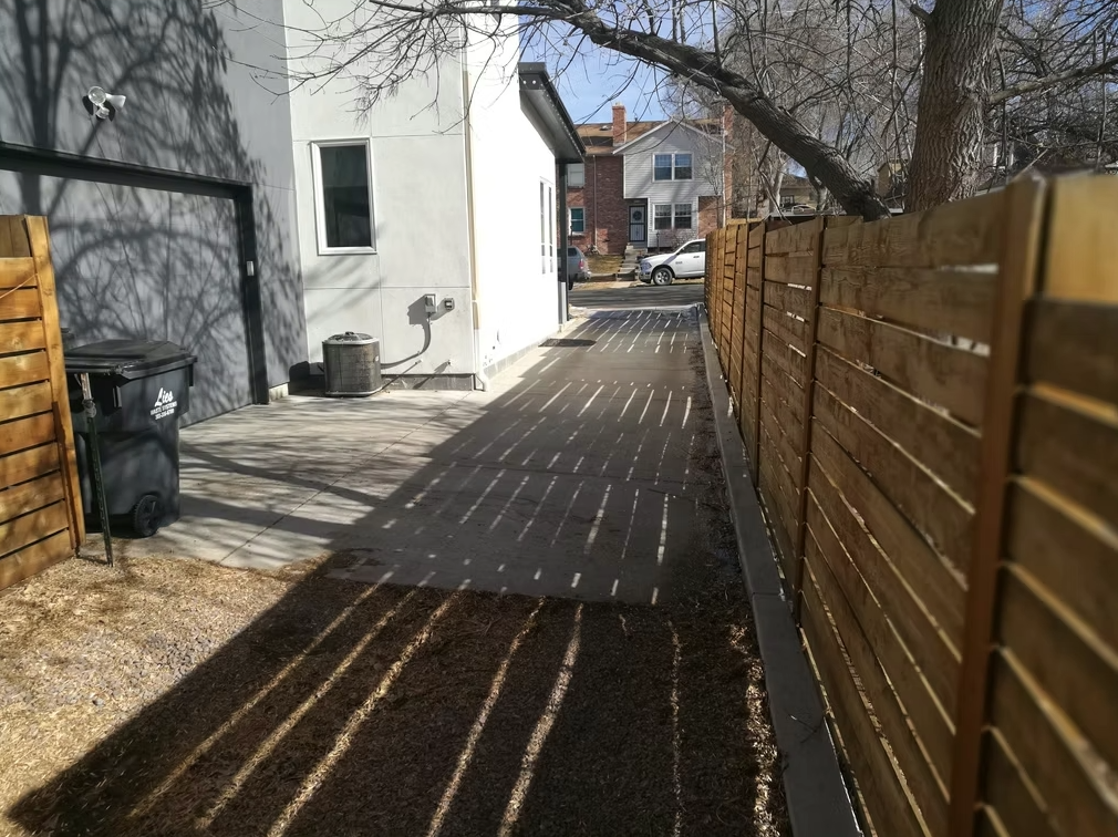 A paved driveway beside a white house is bordered by a wooden fence and bare trees, with stripes of sunlight and shadow on the ground. A black trash bin and parked cars are visible in the background.