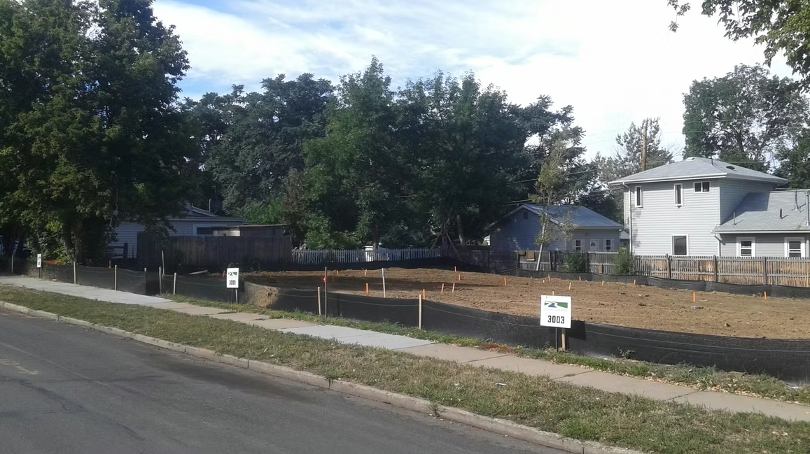 A fenced-off empty lot with signs sits between houses on a residential street. The lot has bare soil and some wooden stakes, with trees and a sidewalk nearby under a partly cloudy sky.