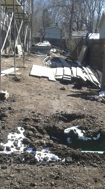 A dirt construction site with scattered building materials, metal scaffolding on the left, some patches of snow, and a fence on the right. Trees and parked cars are visible in the background.