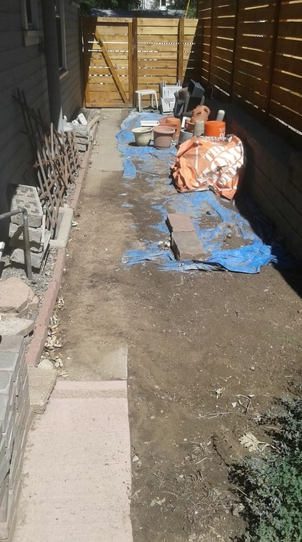 A narrow side yard with a dirt pathway, blue tarp, bricks, pots, and garden tools scattered along a wooden fence, next to a house. A wooden gate and plastic chairs are visible at the end.