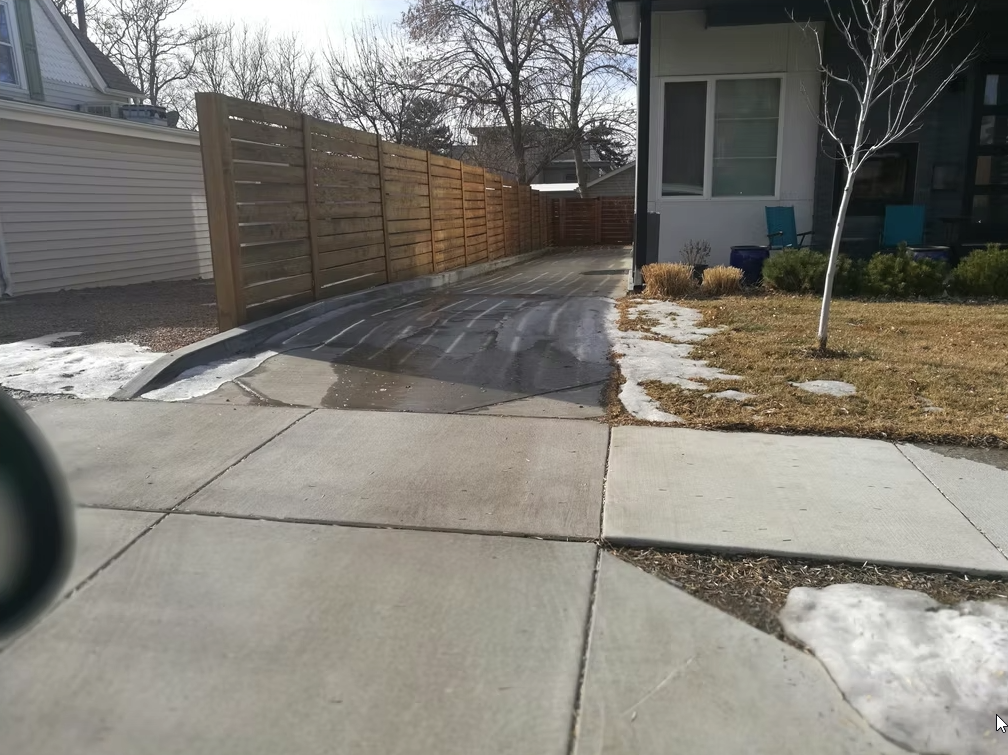A driveway next to a house with patches of melting snow on the grass, wet pavement, a wooden fence, and bare trees under a partly cloudy sky. Two blue chairs are on the porch near the front door.