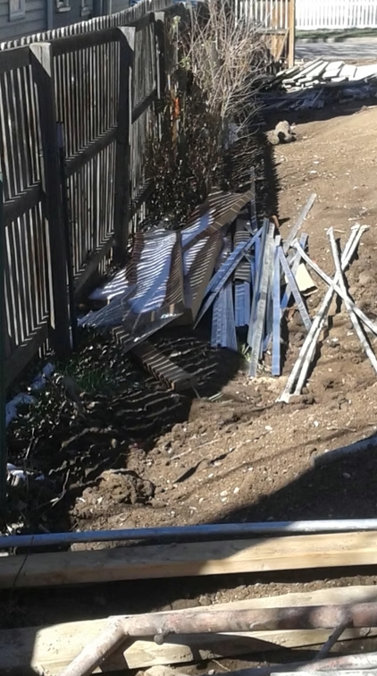 A backyard with dirt ground, scattered metal and wooden planks, and a wooden fence on the left side. Leafless bushes grow near the fence, and construction debris is strewn across the area.