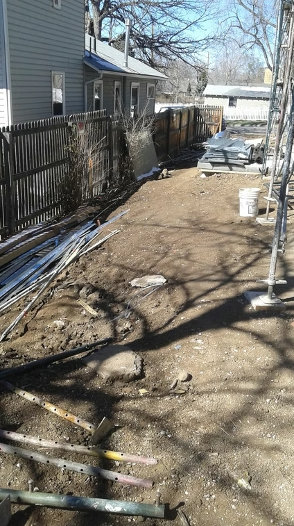 A dirt yard scattered with metal rods, pipes, and construction materials beside a wooden fence, with a gray house on the left and a parked car and trees in the background under clear skies.