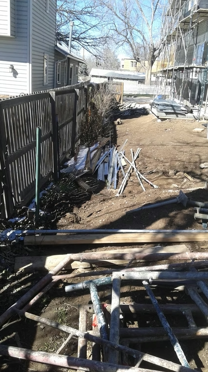 A fenced backyard with scattered construction materials, metal pipes, and wooden planks on bare dirt. A house and scaffolding are visible on the right, with leafless trees in the background under a clear sky.