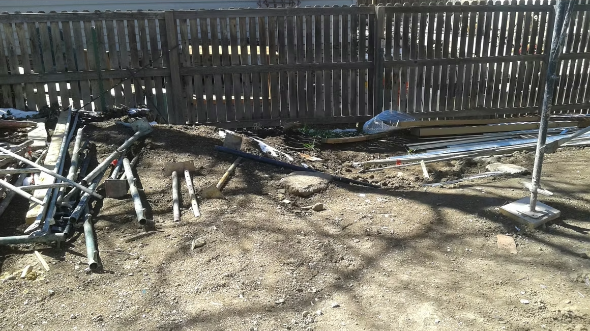 A dirt yard with scattered metal pipes and construction materials, bordered by a wooden fence; some shadows of tree branches are visible on the ground in bright sunlight.