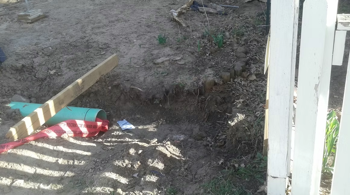 A hole dug in a patch of dirt beside a wooden fence, with a piece of wood, a green and red pipe, and a red caution tape lying nearby; small plants are sprouting in the background.