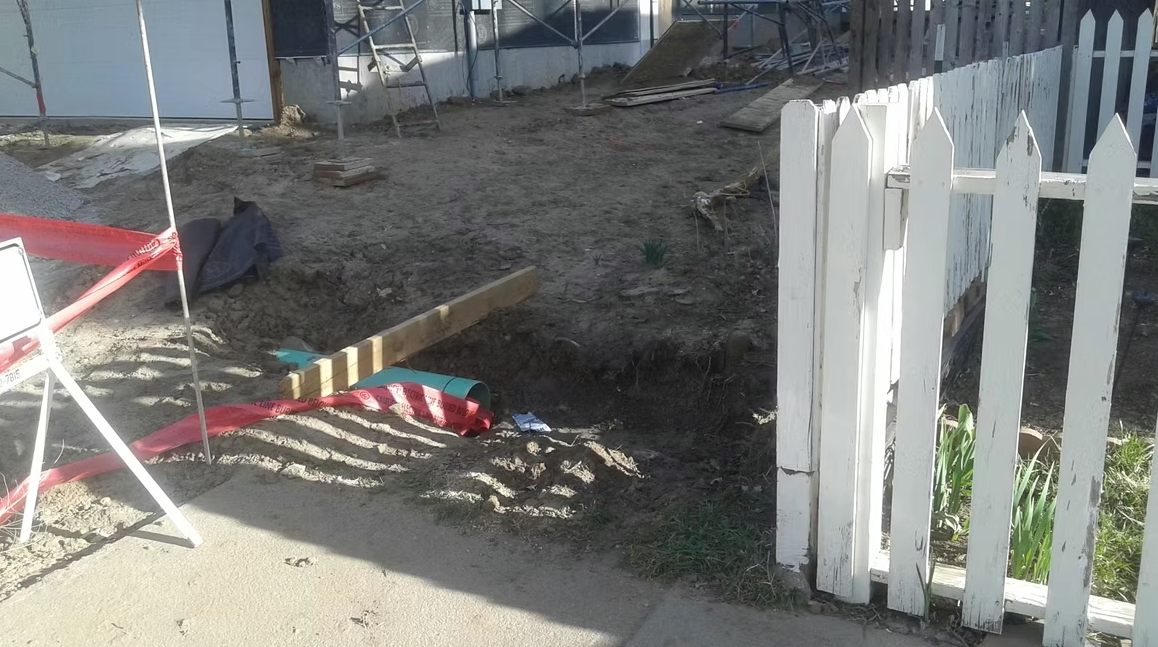 A construction area with a dirt ground, scattered materials, a wooden beam, red caution tape, and an open white picket fence gate. Shadows fall across the sidewalk in the foreground.